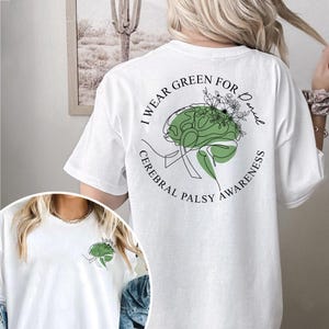 May include: White t-shirt with a green brain graphic and the text "I wear green for Daniel Cerebral Palsy Awareness". The design includes a green brain with floral accents. The shirt is a casual wear item.