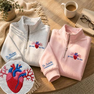 May include: Two quarter-zip sweatshirts in light gray and light pink, each featuring an embroidered anatomical heart with the text "go with the flow" and a sleeve cuff embroidered with "Dr. Marissa CARDIOLOGY" or "Dr. Isabella CARDIOLOGY."