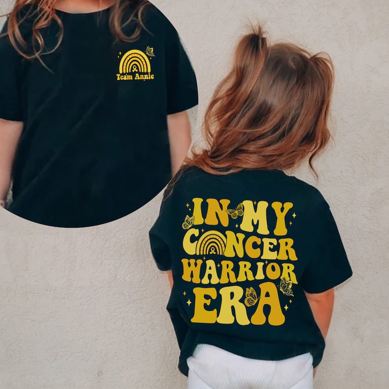 Pu&ograve; includere: Maglietta nera con un design dorato sul retro che dice "In My Cancer Warrior Era" con un arcobaleno e farfalle. La parte anteriore della maglietta ha un design dorato con un arcobaleno e il testo "Team Annie".