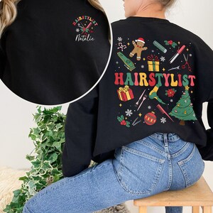 Op de afbeelding: Zwart sweatshirt met een kerstthema ontwerp met een peperkoekman, een kerstboom en de tekst "HAIRSTYLIST". Het sweatshirt heeft ook een kleiner ontwerp aan de voorkant met de tekst "HAIRSTYLIST Natalie".