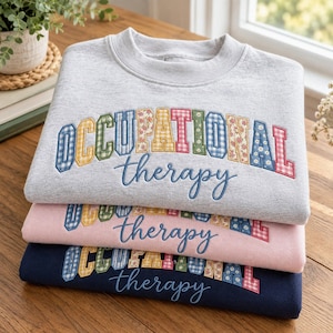 May include: A stack of three crewneck sweatshirts in light gray, light pink, and navy blue. Each features the text OCCUPATIONAL in colorful, patterned block letters and the word therapy in blue cursive script embroidered across the chest.