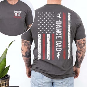 May include: A dark gray t-shirt with a distressed American flag design on the back. The flag has red and white stripes and white stars. The text "Dance Dad" is written vertically down the flag with a silhouette of a dancer. The front of the shirt has the text "Dance D.D. Remarkable" in white.
