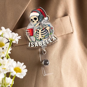 May include: A clear badge reel featuring a skeleton wearing a Santa hat and headset, holding a red cup, and decorated with Christmas lights. The name "ISABELLA" is printed on the badge. The reel is attached to a clear plastic clip.