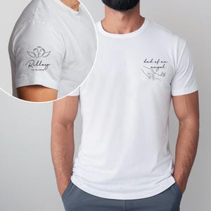 May include: White t-shirt with black text and graphics. The left sleeve features a design with angel wings, baby feet, the name "Riley", and the date "10.15.2024". The front of the shirt says "dad of an angel" with a line drawing of hands.