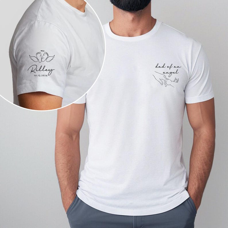 May include: White t-shirt with black text and graphics. The left sleeve features a design with angel wings, baby feet, the name "Riley", and the date "10.15.2024". The front of the shirt says "dad of an angel" with a line drawing of hands.