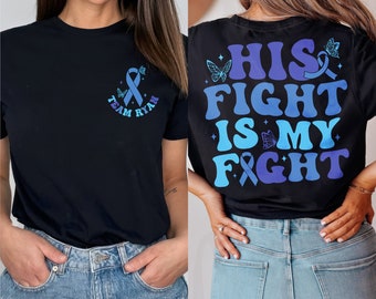 Op de afbeelding: Twee zwarte T-shirts met een blauw lintontwerp. Het voorste shirt heeft de tekst "Team Ryan" en het achterste shirt heeft de tekst "His Fight Is My Fight".