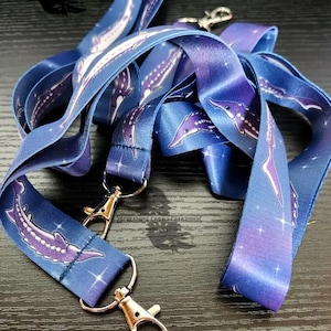 Puede incluir: Un juego de cintas de cordón azul y morado con un diseño de esturión blanco y cierres metálicos plateados. Las cintas tienen un patrón de galaxia con estrellas blancas.