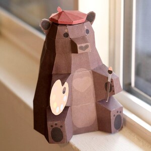 May include: A brown paper bear wearing a red beret holds a paintbrush and a colorful paint palette. The bear is sitting on a windowsill.