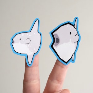 May include: Two finger puppets of a mola mola, a type of fish, with a light gray body and black eyes. The puppets are made of white paper with a blue outline.