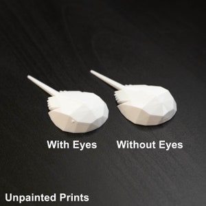 May include: Two white 3D printed horseshoe crab models. One model has eyes and the other does not. The models are on a dark wood surface. Unpainted Prints.