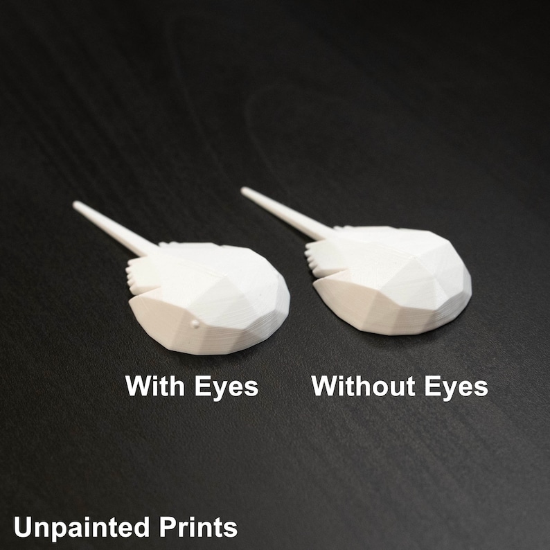 May include: Two white 3D printed horseshoe crab models. One model has eyes and the other does not. The models are on a dark wood surface. Unpainted Prints.