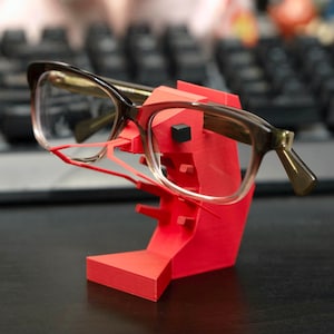 May include: A pair of eyeglasses with brown frames rests on a bright red glasses stand. The stand is 3D-printed and has a geometric design. The background includes a keyboard and a blurred orange object.