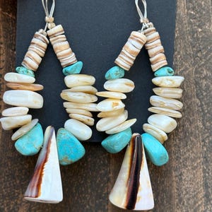 May include: A pair of teardrop-shaped dangle earrings featuring a mix of white shell discs, smooth white stone beads, and bright turquoise stones. Each earring is finished with a large, conical white shell pendant with a dark brown vertical stripe.