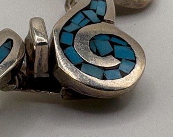 May include: Close-up of a silver-toned jewelry piece with a turquoise inlay. The design features a swirling, abstract shape filled with small, irregular pieces of bright blue stone. The metal has a slightly aged appearance.