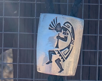 Op de afbeelding: Een zilveren bolo tie met een zwart leren koord heeft een gestileerd ontwerp van een indiaanse danser. Het ontwerp is geëtst in het zilver en toont een figuur met een hoofdtooi en een fluit.