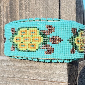 Pode incluir: Uma pulseira de punho turquesa com um padrão repetido de desenhos estilizados de tartarugas em amarelo, laranja e castanho. A pulseira é feita de pequenas contas coloridas e é exibida em uma superfície de madeira.