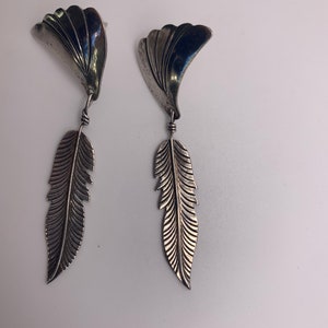 May include: A pair of silver earrings with a feather design. The earrings are made of metal and have a silver finish. The feathers are detailed and have a textured surface.