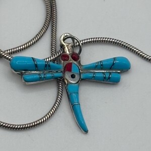 May include: A silver necklace featuring a dragonfly pendant. The pendant has turquoise blue wings, a turquoise body, and red and white accents. The necklace chain is a silver snake chain.
