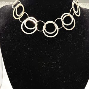 May include: Silver-toned necklace featuring a series of interconnected, open circles of varying sizes. The necklace is displayed on a black mannequin, showcasing its design and structure. The circles are arranged to create a modern, eye-catching accessory.