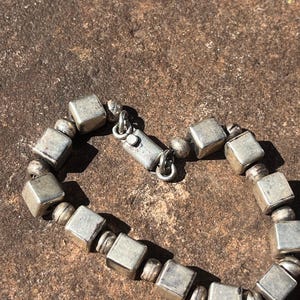 May include: Silver-toned bracelet featuring square and round beads. The bracelet has a clasp closure and is displayed on a textured, brown surface. The beads have a slightly aged appearance.