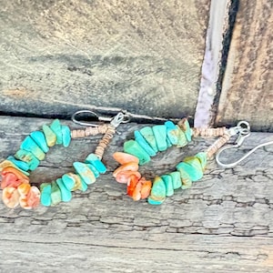 May include: A pair of turquoise and coral chip earrings with silver-colored hooks. The earrings are made of small, irregularly shaped turquoise and coral chips strung together. The earrings are set against a weathered wood background.