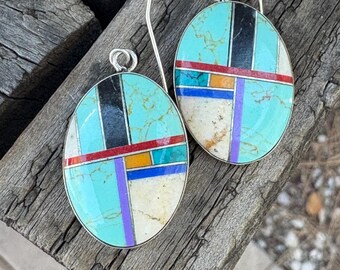 May include: Oval turquoise earrings with inlaid geometric patterns in black, red, orange, purple, and white. The earrings have a silver metal frame and ear wires. The earrings are displayed on a weathered wooden surface.