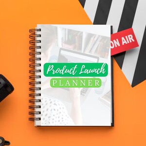 Könnte beinhalten: Ein Spiralnotizbuch mit den Worten "Product Launch PLANNER" in Grün und Weiß. Ein rotes "ON AIR"-Schild und ein schwarzes Mikrofon sind auf einem orangefarbenen Hintergrund zu sehen. Das Notizbuch ist geöffnet, mit einer Person, die an einem Laptop arbeitet, sichtbar.