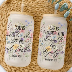 Puede incluir: Dos vasos de vidrio esmerilado con tapas y pajitas de bambú. Cada vaso está decorado con diseños florales y citas inspiradoras. Uno dice "GOD IS within her SHE WILL not fall" y el otro dice "SHE IS CLOTHED WITH strength AND dignity."