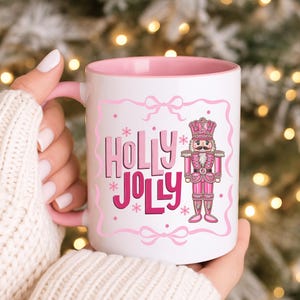 May include: A white ceramic mug with a pink interior and handle. The mug features the text "HOLLY JOLLY" in pink, along with a pink nutcracker illustration. The design is framed with pink bows and snowflakes.
