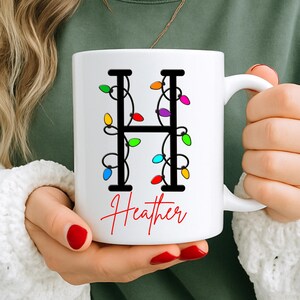 May include: White ceramic mug with a black letter "H" adorned with colourful Christmas lights. The name "Heather" is written in red script below the letter. The mug has a white handle.