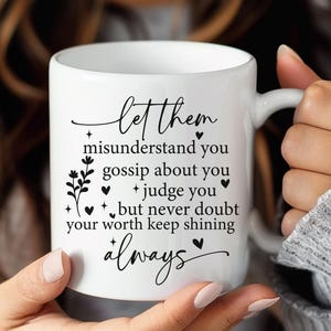May include: White ceramic mug with a black script that reads "Let them misunderstand you, gossip about you, judge you, but never doubt your worth keep shining always." The mug has a handle and is held by a person.