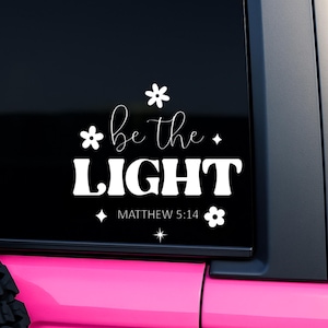 May include: A white decal on a black background with the words "be the LIGHT" and the reference "MATTHEW 5:14". The decal is decorated with floral and star designs. The decal is on the rear window of a pink vehicle.