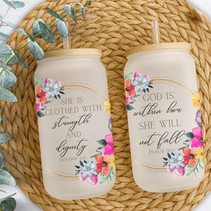 May include: Two frosted glass tumblers with bamboo lids and straws. Each tumbler features floral designs in pink, blue, and yellow, with gold accents. One reads "She is clothed with strength and dignity," the other "God is within her, she will not fall."