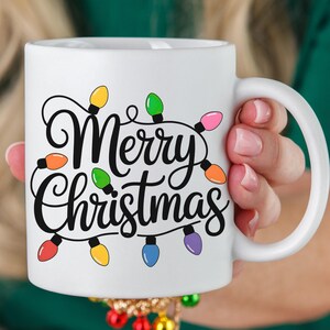 May include: White ceramic mug with the words "Merry Christmas" in black script, adorned with colorful Christmas lights. The mug has a white handle and is held by a person.