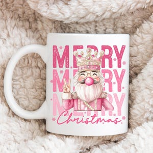 May include: White ceramic mug featuring a pink illustration of a Santa Claus-like figure wearing a crown and glasses, giving a peace sign. The mug has the words "MERRY Christmas" in pink.