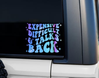 May include: A vinyl sticker with the text "EXPENSIVE DIFFICULT & TALKS BACK" in a retro font. The text is in shades of blue, purple, and pink, with star accents. The sticker is on a car window.