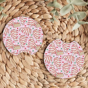 May include: Two car coasters with a pink and white checkered background. The coasters feature a pattern of red and white striped Christmas trees, cake slices, and green and red stars.