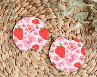 May include: Two pink and white car coasters with a strawberry and flower pattern. The coasters are round and have a cut-out for a cup.