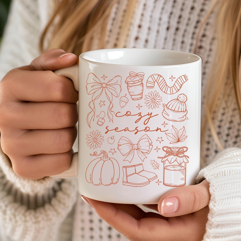 May include: White ceramic mug with a beige handle, featuring a "cozy season" design in orange. The design includes bows, a pumpkin, a scarf, a hat, and other fall-themed illustrations. The mug is held by a person.