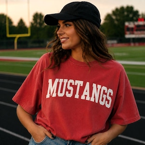 May include: A person wearing a red t-shirt with the word "MUSTANGS" in white block letters and a black baseball cap. The person is standing on a track field with a football field in the background.