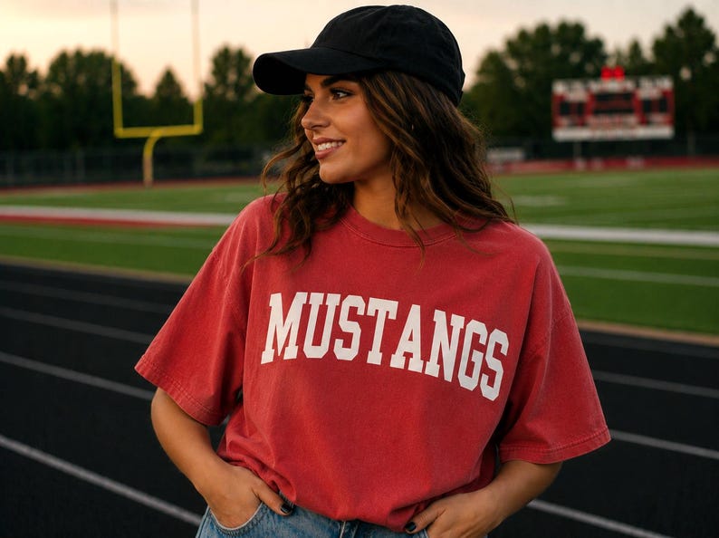 May include: A person wearing a red t-shirt with the word "MUSTANGS" in white block letters and a black baseball cap. The person is standing on a track field with a football field in the background.