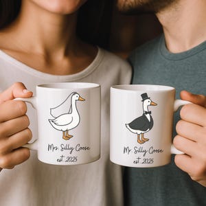 Puede incluir: Dos tazas de cer&aacute;mica blanca con asas negras. La taza de la izquierda tiene una ilustraci&oacute;n de dibujos animados de un ganso blanco con velo y el texto "Mrs. Silly Goose est. 2025". La taza de la derecha tiene una ilustraci&oacute;n de dibujos animados de un ganso con esmoquin y el texto "Mr. Silly Goose est. 2025".