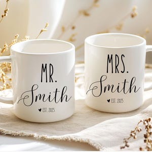 May include: Two white ceramic mugs with black lettering. The mug on the left says "MR. Smith EST. 2025" and the mug on the right says "MRS. Smith EST. 2025".