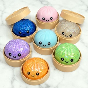 May include: A collection of seven round, squishy dumpling toys, each resting in a small beige plastic steamer basket. The dumplings come in various colors including pink, blue, purple, orange, green, and clear, all featuring glitter and faces.