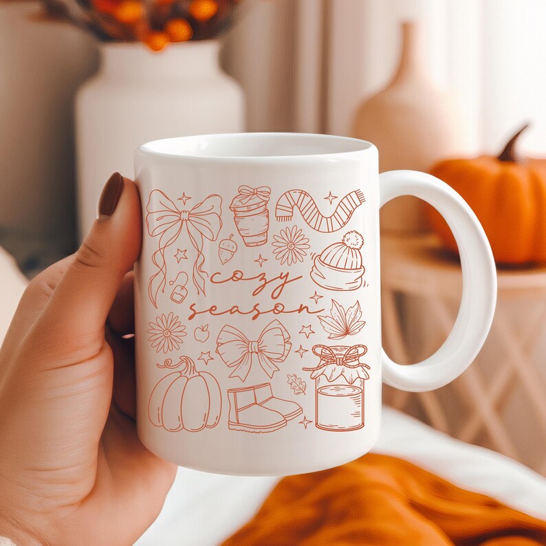 May include: White ceramic mug with a handle, featuring a "cozy season" design in orange. The design includes illustrations of bows, a coffee cup, a scarf, a hat, flowers, a pumpkin, boots, and a jar. The mug is held by a person.