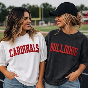 May include: Two women wearing white and charcoal gray t-shirts with the words "CARDINALS" and "BULLDOGS" in red lettering. One woman wears a black baseball cap. Both are smiling, standing on a track field.