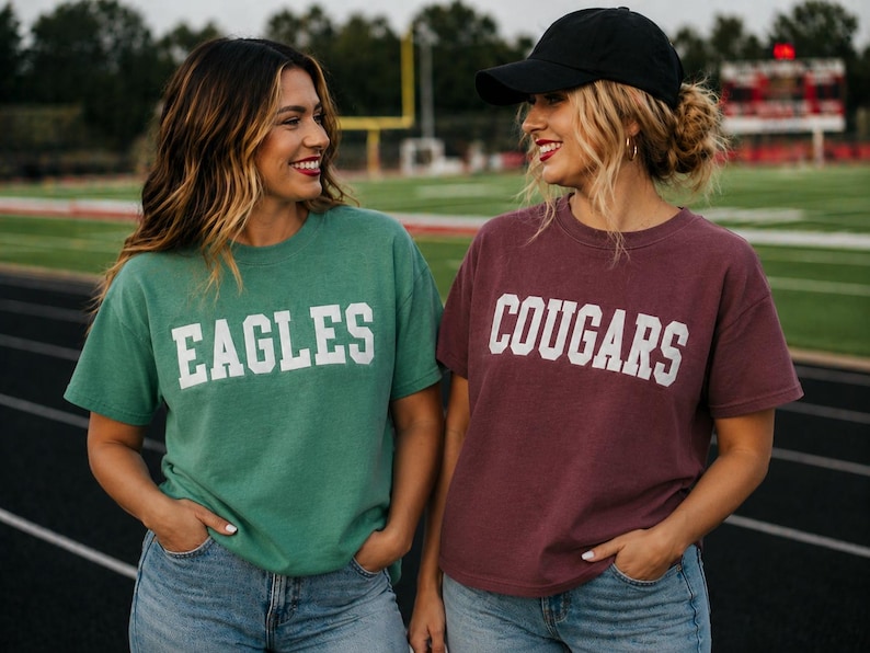 May include: Two people wearing matching t-shirts with team names. One shirt is green with "EAGLES" in white block letters. The other is maroon with "COUGARS" in white block letters. Both are wearing blue jeans.