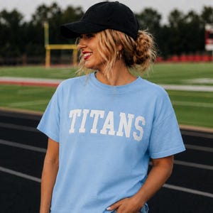 May include: A light blue t-shirt with the word "TITANS" in white block letters. The person in the image is wearing a black baseball cap. The background is a green field and track.