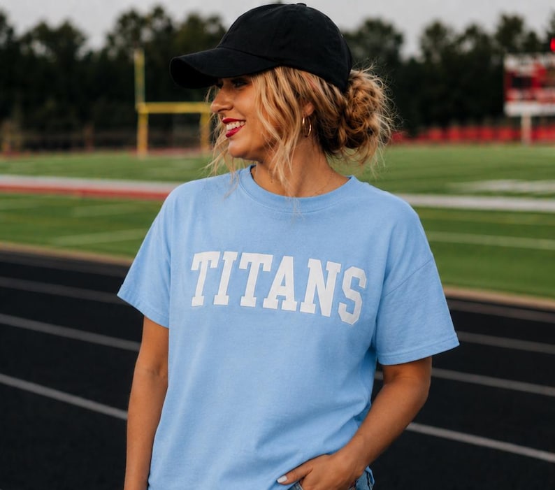 May include: A light blue t-shirt with the word "TITANS" in white block letters. The person in the image is wearing a black baseball cap. The background is a green field and track.
