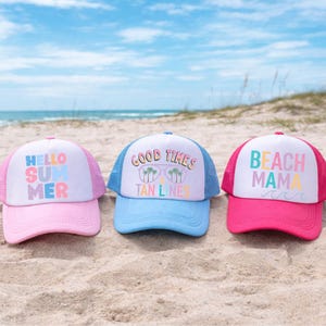 May include: Three colorful trucker hats with white mesh backs and curved brims are displayed on a sandy beach. The hats feature the phrases "HELLO SUMMER", "GOOD TIMES TAN LINES", and "BEACH MAMA" in pastel colors.